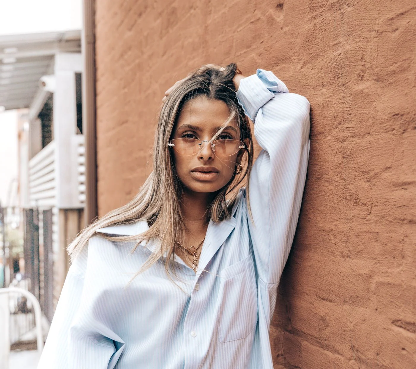 Mulher de óculos e camisa azul junto a uma parede em tom terracota